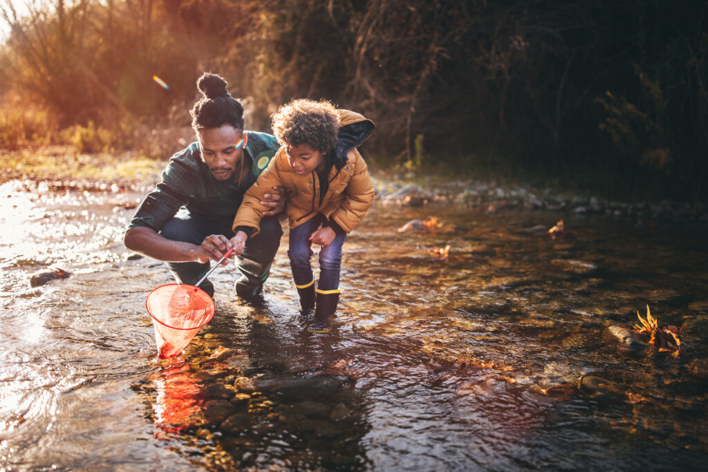 Young dad teaching son how to fish with fishing net in mountain stream at sunset