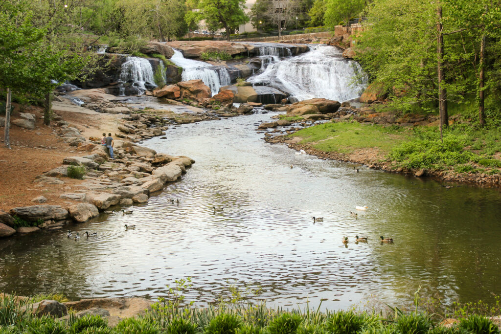 Golden hour view of waterfall and river in Greenville Falls park with young couple looking at the waterfall and ducks swimming in the sunlit river